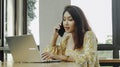 Young Asian woman working on laptop computer while talking on the phone at the cafe, looking through documents Royalty Free Stock Photo