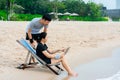 Young Asians sit on a laptop video chatting on a laptop, in a chair by the sea, comfortably, having fun chatting