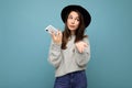 young asking dissatisfied brunette woman wearing black hat and grey sweater holding smartphone looking to the side