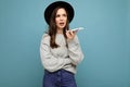 young asking dissatisfied brunette woman wearing black hat and grey sweater holding smartphone looking to the side