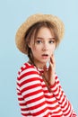 Young astonished girl with two braids in straw hat and red striped vest holding hand near face while thoughtfully