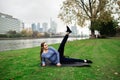 Young athlete woman doing exercises lying on the grass near the river, against city background