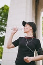 Young athletic beautiful brunette girl in black uniform, cap with headphones holding bottle drinking water during Royalty Free Stock Photo