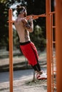 Young athletic man in headband with naked torso dressed in black leggings and red shorts pulls up on the horizontal bar