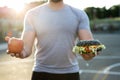 Young athletic man in a t-shirt holds a Burger and an Apple in the background of the stadium, a choice of healthy and Royalty Free Stock Photo