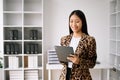 Young attractive Asian female office worker business suits smiling at camera with working notepad, tablet and laptop documents .