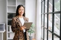 Young attractive Asian female office worker business suits smiling at camera with working notepad, tablet and laptop documents .