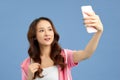 Young attractive Asian girl having video-call phone in hand, shooting selfie on front camera isolated on blue background