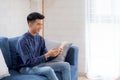 Young attractive asian man resting using browsing tablet computer on sofa at home happy male sitting on couch relax reading Royalty Free Stock Photo