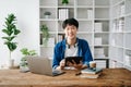 Young attractive Asian man smiling thinking planning writing in notebook, tablet and laptop working from home at home office Royalty Free Stock Photo