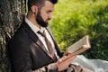 Young attractive bearded business man sitting on green grass under tree and resting in park. Read book, drink coffee Royalty Free Stock Photo