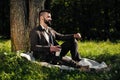 Young attractive bearded business man sitting on green grass under tree and resting in park. Read book, drink coffee Royalty Free Stock Photo