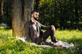 Young attractive bearded business man sitting on green grass under tree and resting in park. Read book, drink coffee Royalty Free Stock Photo