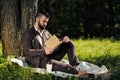 Young attractive bearded business man sitting on green grass under tree and resting in park. Read book, drink coffee Royalty Free Stock Photo