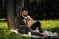 Young attractive bearded business man sitting on green grass under tree and resting in park. Read book, drink coffee Royalty Free Stock Photo