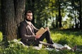 Young attractive bearded business man sitting on green grass under tree and resting in park. Read book, drink coffee Royalty Free Stock Photo