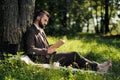 Young attractive bearded business man sitting on green grass under tree and resting in park. Read book, drink coffee Royalty Free Stock Photo