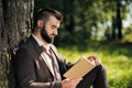 Young attractive bearded business man sitting on green grass under tree and resting in park. Read book, drink coffee Royalty Free Stock Photo