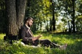 Young attractive bearded business man sitting on green grass under tree and resting in park. Read book, drink coffee Royalty Free Stock Photo