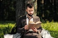 Young attractive bearded business man sitting on green grass under tree and resting in park. Read book, drink coffee Royalty Free Stock Photo