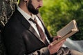 Young attractive bearded business man sitting on green grass under tree and resting in park. Read book, drink coffee Royalty Free Stock Photo