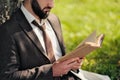 Young attractive bearded business man sitting on green grass under tree and resting in park. Read book, drink coffee Royalty Free Stock Photo