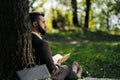 Young attractive bearded business man sitting on green grass under tree and resting in park. Read book, drink coffee Royalty Free Stock Photo