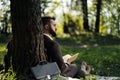 Young attractive bearded business man sitting on green grass under tree and resting in park. Read book, drink coffee Royalty Free Stock Photo