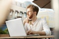 Young attractive bearded ginger guy with earring in white shirt drinks coffee, working on new project remotely on his Royalty Free Stock Photo