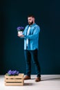 Young attractive bearded hipster man, dressed in denim shirt and blue pants, stands indoors, holding pot of flower and looking at Royalty Free Stock Photo