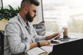 Young attractive bearded hipster man sits in cafe in front of aptop, reading paper documents. Freelancer works remotely