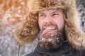 A young attractive bearded man in a fur hat and with snow on his beard shows his tongue and looks to the side. Tries to