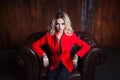 Young and attractive blond woman in red jacket sits in leather armchair, background grunge rusty wall Royalty Free Stock Photo