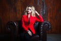 Young and attractive blond woman in red jacket sits in leather armchair, background grunge rusty wall Royalty Free Stock Photo