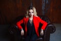 Young and attractive blond woman in red jacket sits in leather armchair, background grunge rusty wall Royalty Free Stock Photo