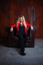 Young and attractive blond woman in red jacket sits in leather armchair, background grunge rusty wall Royalty Free Stock Photo