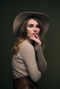 A young attractive blonde woman in a beige sweater, fedora hat and brown leather trousers poses on a dark green olive Royalty Free Stock Photo