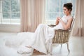 Bride in beautiful white dress sitting on chair indoors in white studio interior like at home.