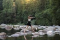 Young attractive brunette woman hopping over rocks beside a pristine river in an evergreen forest.