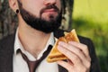 Young attractive business man with beard sitting on green grass under tree and resting in park. Bites sandwich close up Royalty Free Stock Photo