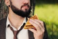 Young attractive business man with beard sitting on green grass under tree and resting in park. Bites sandwich close up Royalty Free Stock Photo