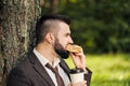 Young attractive business man with beard sitting on green grass under tree and resting in park. Bites sandwich close up Royalty Free Stock Photo