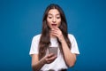 Young and attractive caucasian or arab brunette girl in white t-shirt shocked using mobile phone isolated on blue studio backgroun