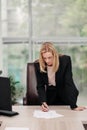 Young attractive Caucasian blond woman in black business suit sits at desk in bright office. Studying paper documents Royalty Free Stock Photo