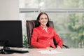 A young attractive Caucasian blond woman in a red business suit sits at a desk in a bright office. Portrait of a Royalty Free Stock Photo