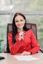 A young attractive Caucasian blond woman in a red business suit sits at a desk in a bright office. Portrait of a Royalty Free Stock Photo