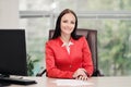 A young attractive Caucasian blond woman in a red business suit sits at a desk in a bright office. Portrait of a Royalty Free Stock Photo