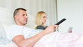 Young attractive caucasian couple in bed. Man watching tv, woman browsing web on digital tablet. Couple in bed not speaking