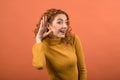 Young and attractive caucasian redhead girl holding her hand near her ear due to deafness isolated on orange studio background. Royalty Free Stock Photo