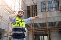 Young attractive construction smiling bearded using smartphone calling to client, worker in vest with white helmet standing on